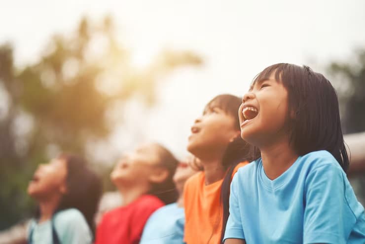 Four children outdoors in a row, heads tipped back, laughing together in warm afternoon light